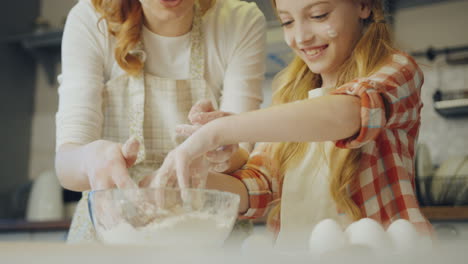 Happy-young-mother-showing-her-teen-blonde-daughter-how-to-kneading-a-daugh-in-a-bowl-while-they-making-food-in-the-kitchen.-Close-up.-Indoor